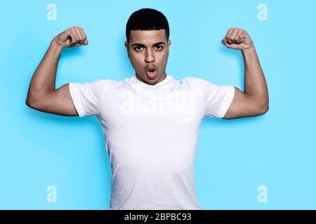 Young strong man in white t-shirt with arms raised showing her biceps while looking camera against blue background Stock Photo