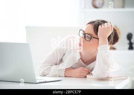 An upset young woman is sitting in an office at a laptop, and is holding her head in her hands. Problems at work Stock Photo