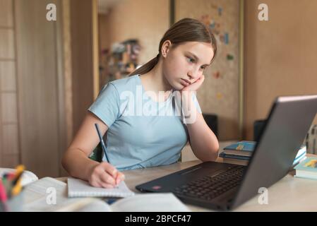 Tired girl is a teenager of 12-15 years old, writes with pen at home, writes down blackboard lessons, learns on Internet, laptop with Internet Stock Photo