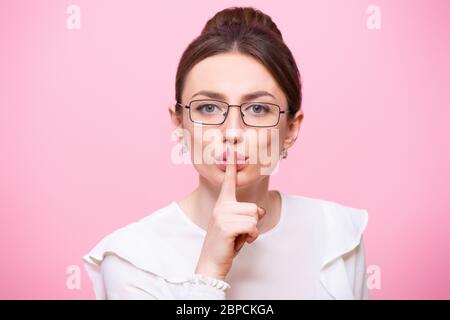 Young woman in glasses holds an index finger near mouth, calling for silence. Hush gesture, quieter. Close-up portrait isolated on a pink background Stock Photo