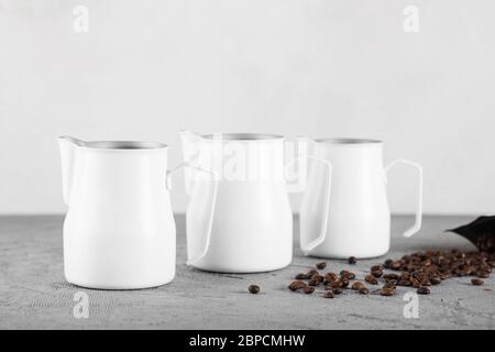 Three white milk jugs on a gray decorative background. Coffee beans are scattered nearby. Stock Photo