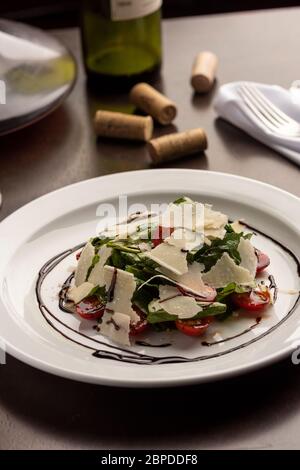 Arugula, tomatoes and parmesan salad dressed with balsamic vinegar on a wooden table, napkin, wine bottle glass and corks. Stock Photo