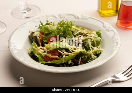 Green salad with lettuce tomatoes on white blurred background with wine glasses, vinegar and olive oil. Stock Photo