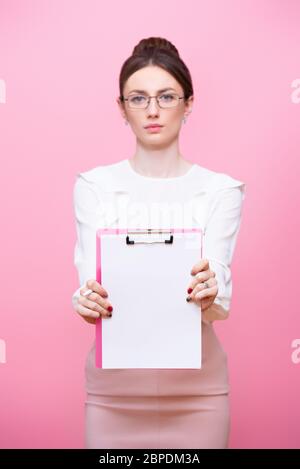 A young woman with glasses holds a clipboard folder in her hands with a serious expression on her face. Isolated on pink background Stock Photo