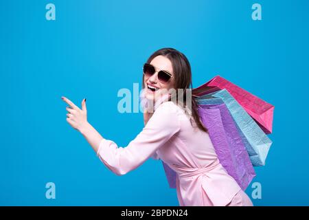 A fashionably dressed girl in sunglasses with multi-colored shopping bags is happy with the discounts and sale. Isolated on a blue background Stock Photo