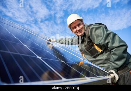 Cheerful male worker smiling to the camera while installing blue photovoltaic solar panel. Electrician in safety helmet under beautiful sky. Concept of alternative energy sources and innovations. Stock Photo