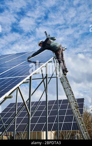 Vertical snapshot of working process in a solar field. Technician in protective uniform, standing on a ladder checking solar batteries on metal ground mounting. Concept of renewable energy resource Stock Photo