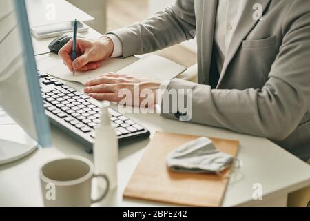 Close-up of businessman sitting at desk with sanitizer and mask and making notes in notepad while working in office during coronavirus quarantine Stock Photo