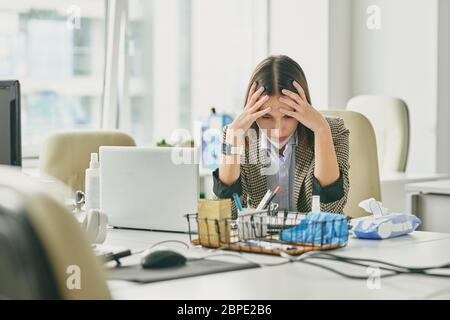 Puzzled young office woman sitting at desk in empty office during coronavirus epidemic and holding head in hands while having problems Stock Photo