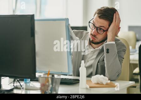 Irritated young businessman in eyeglasses sitting at desk and looking at computer screen while having work problems because of coronavirus quarantine Stock Photo