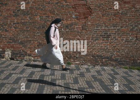 A nun with face mask walking downhill from the Prague Castle with black backpack, large brick wall in the background, no other people in the image Stock Photo