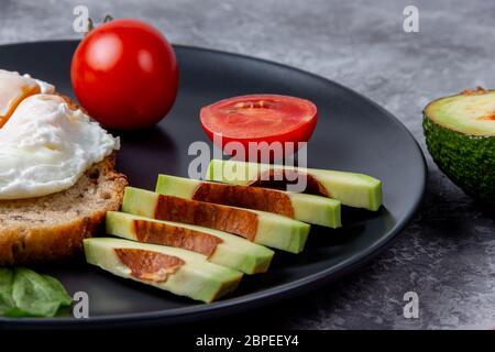 Poached egg with avocado on whole grain bread close to red cherry and spinach on black plate Stock Photo