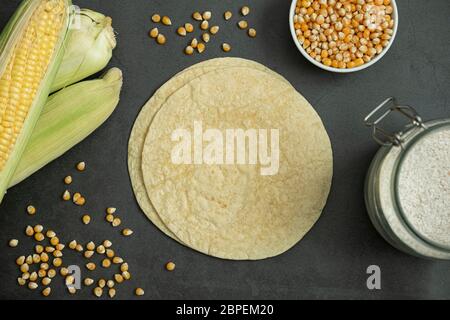 Fresh baked tortilla bread, corn flour in jar, corn kernel in bowl and green corn cobs on concrete, stone background. Delicious, traditional, healthy Stock Photo
