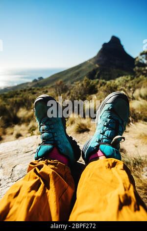 Feet of a female mountaineer relaxing on top of the cliff. Personal perspective of woman hiker resting on mountain trail. Stock Photo