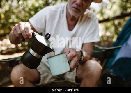 Close-up of a senior man pouring coffee in a mug at campsite. Male traveller having coffee at camping trip. Stock Photo