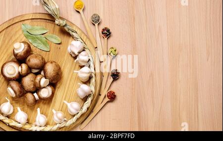 Braid of garlic, mushrooms and bay leaves on a round cutting board light wooden table. Nearby are several small wooden spoons with different spices. T Stock Photo