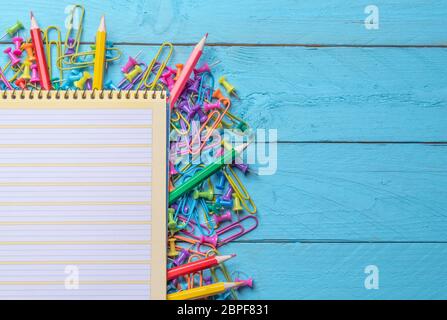 Spiral notebook on a pile of office objects, colored paper clips, crayons and push pins on a blue wooden table Stock Photo