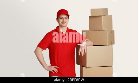 Smiling courier stands in storage warehouse and puts hand on boxes, studio shot Stock Photo