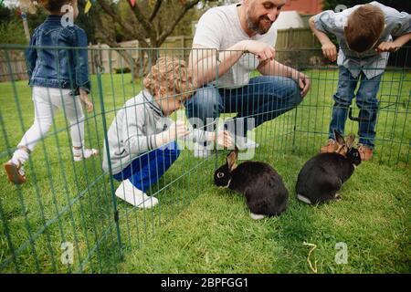 Group of children kneeling outdoors next to a rabbit pen. They are trying to pet the rabbits by putting their fingers through the holes in the fence. Stock Photo