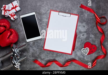 Dumbbells, boxing gloves, gift boxes, red hearts, computer tablet and tablet with blank sheet on grey background.Top view. Valentine's Day card. Fitne Stock Photo
