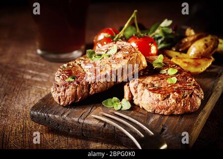 Thick tender roasted or grilled beef fillet medallions served on an old vintage wooden board with tomatoes and potato wedges Stock Photo