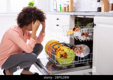 Worried Young Woman Looking At The Dirty Colorful Plates Arranged In The Dishwasher Stock Photo