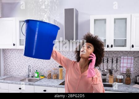 Worried Young Woman Calling Plumber On Cellphone And Collecting Leakage Water From Ceiling Stock Photo