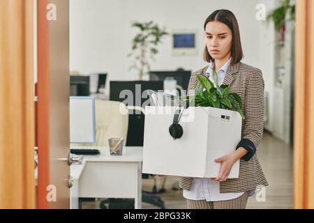 Sad young woman carrying box full of personal stuff while leaving office after retrenchment in coronavirus crisis Stock Photo