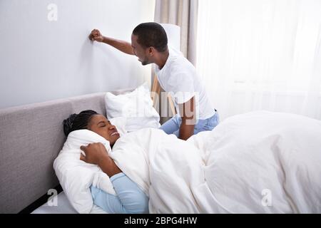 Woman Covering Her Ears With Pillow And Husband Banging Wall For Being Disturbed By Noise Stock Photo