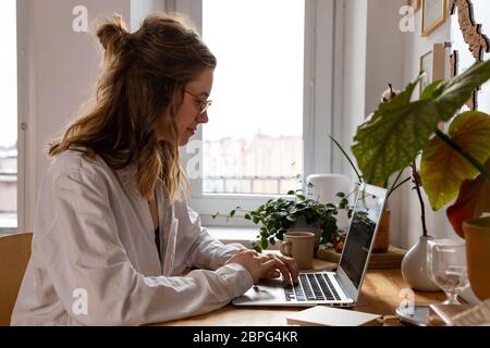 Young woman freelancer/designer working on computer from home office during self-isolation due to coronavirus. Cozy workplace surrounded by plants. Re Stock Photo