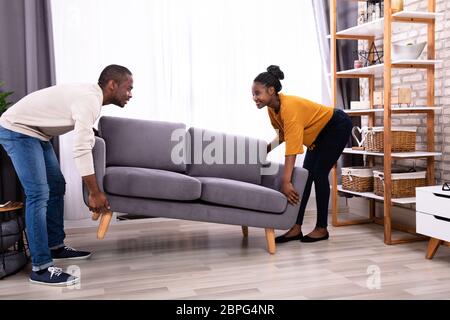 Side View Of A Smiling Young African Couple Lifting Sofa In Living Room Stock Photo