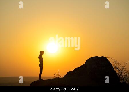 Silhouette of a woman hiker standing alone near big stone at sunset in mountains. Female tourist on high rock in evening nature. Tourism, traveling an Stock Photo