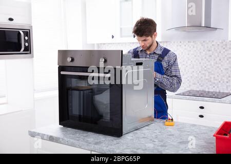 Young Male Repairman Repairing Oven Using Digital Multimeter In Kitchen Stock Photo