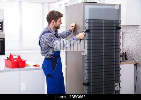 Serviceman In Overall Working On Fridge With Wrench In Kitchen Stock Photo