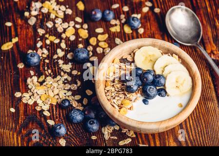Bowl of Granola, Banana, Blueberry and Greek Yoghurt. Scattered Ingredients and Spoon on Wooden Table. Stock Photo