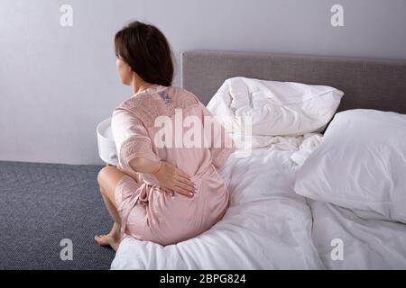 Rear View Of A Woman Sitting On Bed Suffering From Back Pain In Bedroom Stock Photo
