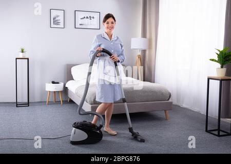Happy Young Housekeeper Cleaning Carpet With Vacuum Cleaner In Hotel Room Stock Photo