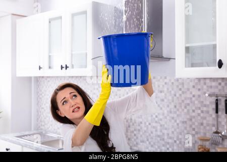 Unhappy Young Woman Collecting Water Leaking From Ceiling In Blue Bucket Stock Photo