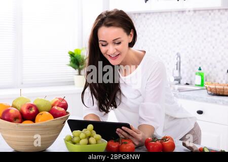 Happy Young Woman Using Digital Tablet Near Fresh Vegetables On Kitchen Counter Stock Photo