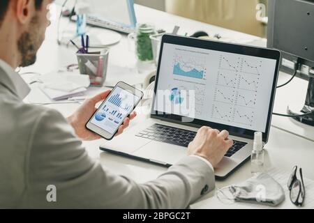Over shoulder view of businessman using smartphone and laptop while analyzing sales graphs during quarantine Stock Photo
