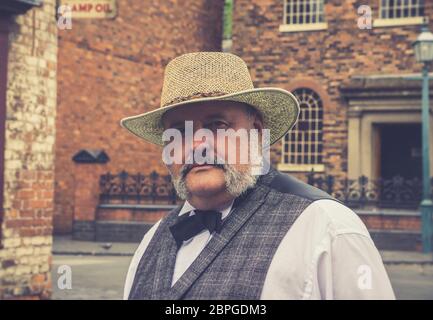 Retro, close-up portrait of smartly-dressed man with moustache in straw hat, isolated outdoors in vintage street at Black Country Living Museum, UK. Stock Photo