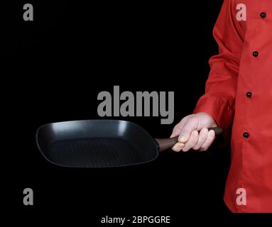 cook in red uniform holding an empty square black frying pan, black background Stock Photo