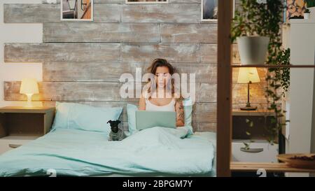 Beautiful caucasian woman in pajamas browsing on laptop laying on bed with her dog.
