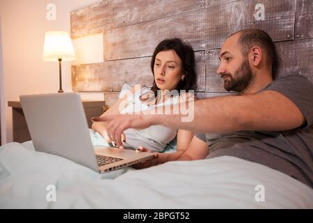 Husband pointing at laptop before bedtime. Happy married couple in pajamas. Stock Photo