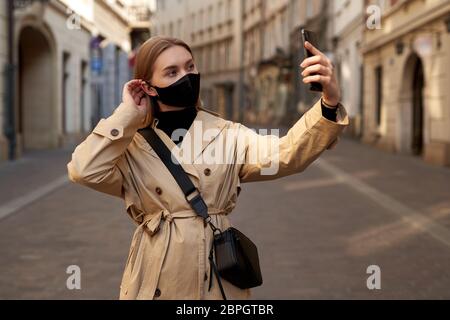 Blone Young attractive female traveler makes selfie, wears protective medical mask to protect herself from coronavirus. Pandemic concept Stock Photo