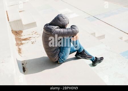 a young caucasian man, wearing jeans and a gray hooded jacket, curled up sitting on an outdoor concrete stairway Stock Photo