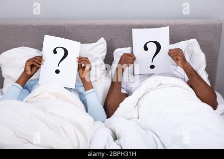 Couple Lying On Bed Holding Question Mark Sign In Front Of Their Face Stock Photo