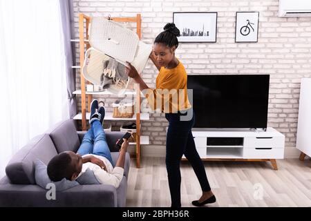 Young Woman Throwing Dirty Clothes On Lazy Husband Lying On Sofa Stock Photo