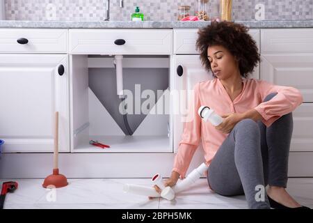 Disappointed African Young Woman Holding Broken Sink Pipe In Hand Sitting On The Kitchen Floor Stock Photo