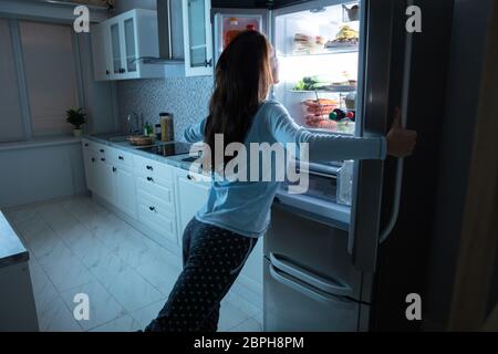 Woman Opening Refrigerator Door With Various Food In Kitchen Stock Photo